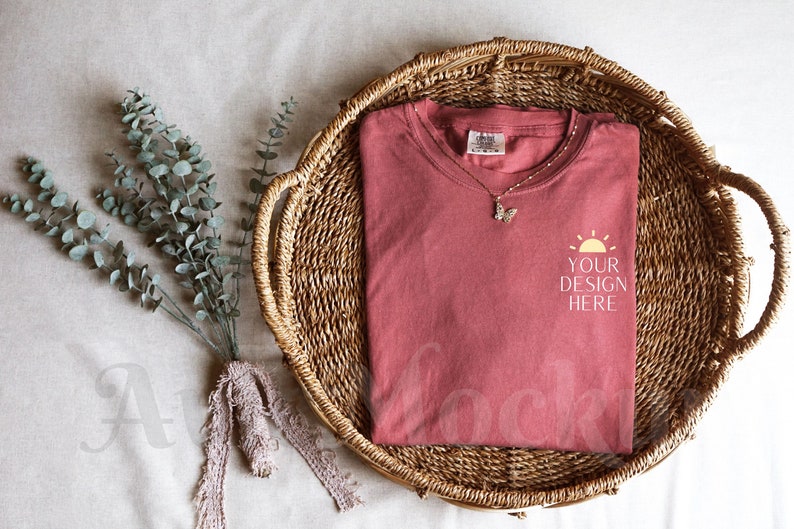 K&ouml;nnte beinhalten: Ein rotes T-Shirt mit einem wei&szlig;en Sonnenmotiv und dem Text "YOUR DESIGN HERE" auf einem Weidenkorb.