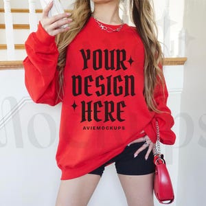 May include: Red sweatshirt with long sleeves and a crew neck. The sweatshirt has the text "Your design here" in black gothic font. The text "Aviemockups" is printed in black below the main text.