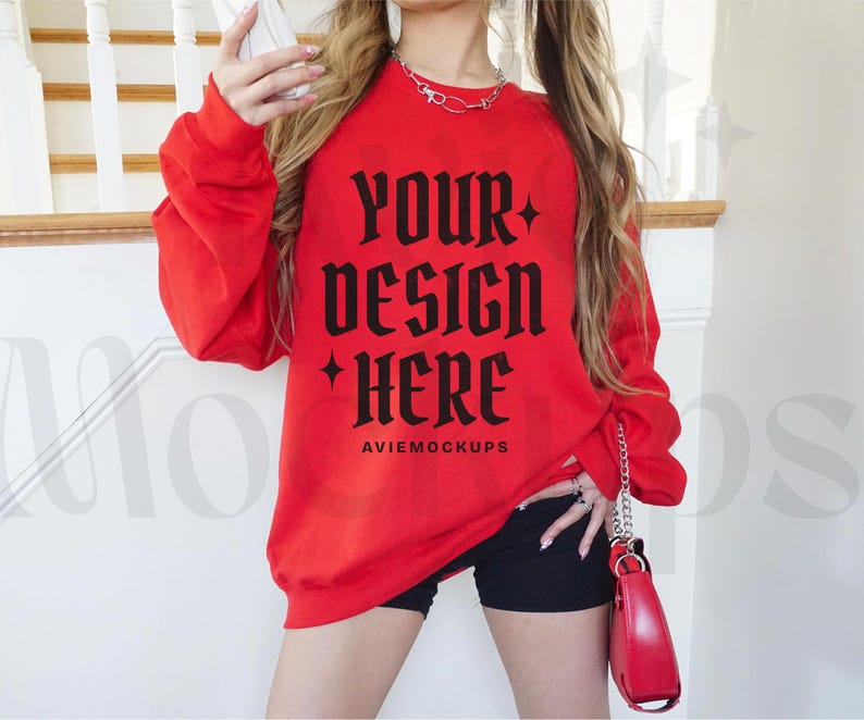 May include: Red sweatshirt with long sleeves and a crew neck. The sweatshirt has the text "Your design here" in black gothic font. The text "Aviemockups" is printed in black below the main text.