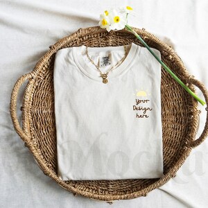 May include: A white t-shirt with a simple design of a sun and the text "Your Design Here" is displayed on a woven basket with a white flower.