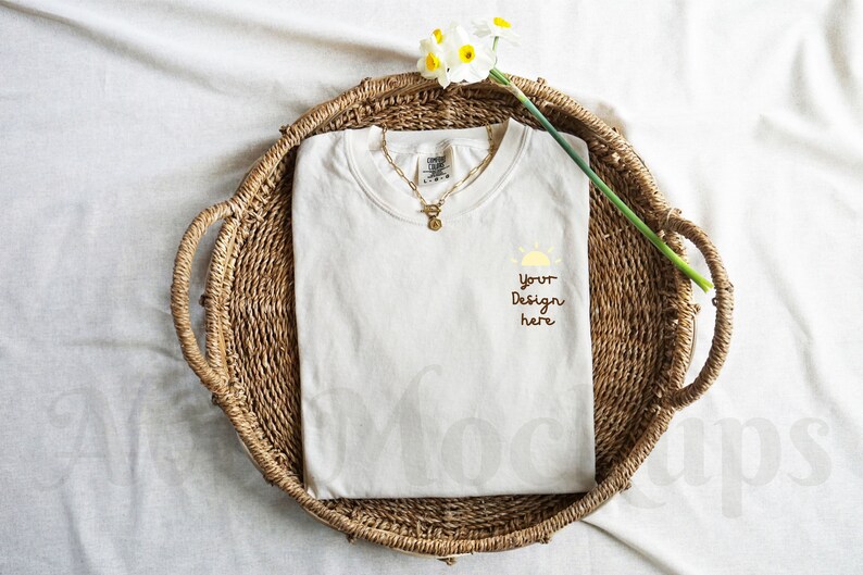 May include: A white t-shirt with a simple design of a sun and the text "Your Design Here" is displayed on a woven basket with a white flower.