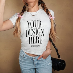 Może przedstawiać: Biała koszulka z napisem "YOUR DESIGN HERE" i słowami "AVIEMOCKUPS" poniżej. Koszulkę nosi osoba z długimi blond włosami zaplecionymi w warkocze z różowymi wstążkami. Osoba nosi niebieskie dżinsy i czarną torbę.