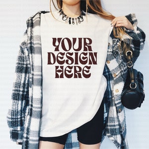 以下が含まれることがあります： 白いTシャツに、茶色の文字で「YOUR DESIGN HERE」と書かれています。シャツは、長い茶色の髪とチェックシャツを着た人が着ています。