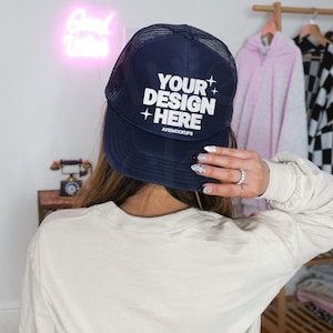 May include: Navy blue trucker hat with white text that reads "YOUR DESIGN HERE" and the website "AVIEMOCKUPS". The hat has a mesh back and a curved brim. The hat is being held up by a person.