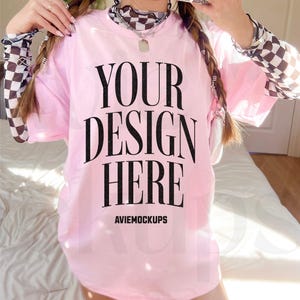 May include: A pink oversized t-shirt with the text "YOUR DESIGN HERE" and the text "AVIEMOCKUPS" below it. The t-shirt is being worn by a person with long braided hair and a checkered shirt underneath.