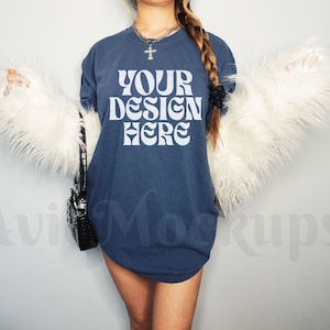Pode incluir: Uma camiseta azul oversized com o texto "YOUR DESIGN HERE" em branco. A camiseta está sendo modelada por uma pessoa com cabelo castanho longo e um colar preto.