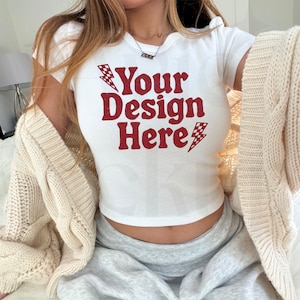 May include: White crop top with the text "Your Design Here" in red, with lightning bolt graphics. A cream-coloured cardigan is draped over the shoulders. The model is wearing grey sweatpants.