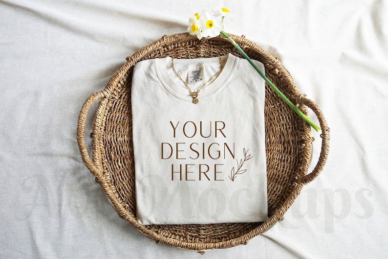May include: White t-shirt with the text "YOUR DESIGN HERE" and a small leaf design, laying in a woven basket with a white flower stem.