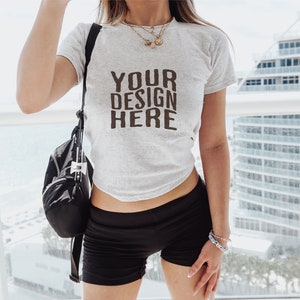 Könnte beinhalten: Eine Frau trägt ein weißes T-Shirt mit dem Aufdruck "YOUR DESIGN HERE". Sie trägt auch schwarze Shorts und einen schwarzen Rucksack.
