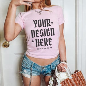 Könnte beinhalten: Ein rosa, bauchfreies T-Shirt mit dem Text "YOUR DESIGN HERE" und den Worten "AVIEMOCKUPS" darunter. Das Shirt wird von einer Person mit Jeans-Shorts und einer Strohtasche getragen.
