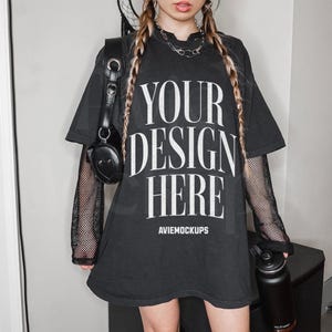 May include: Black oversized t-shirt with the text "YOUR DESIGN HERE" and the words "Aviemockups" printed below. The t-shirt has long sleeves with a black mesh overlay. The model is wearing a black choker necklace and has long blonde braids.