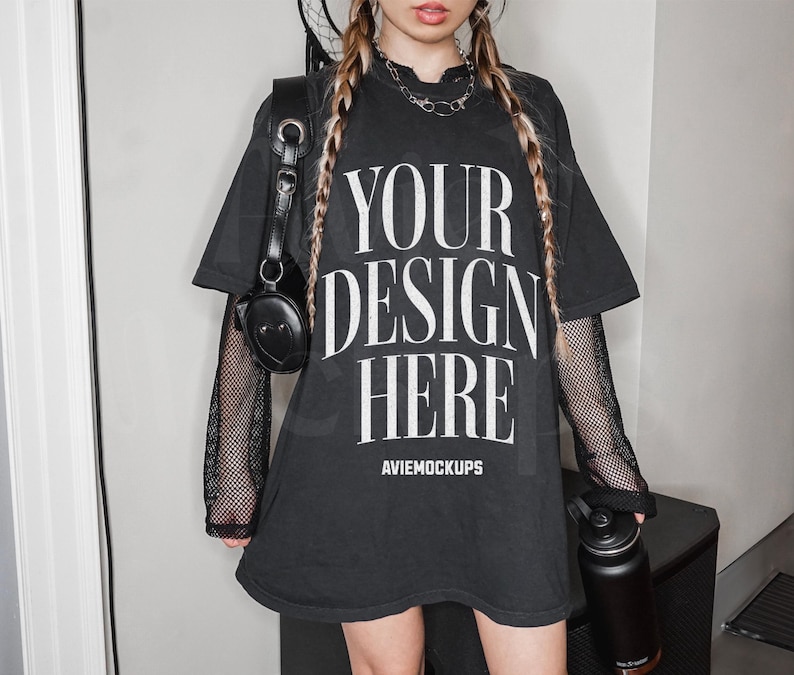 May include: Black oversized t-shirt with the text "YOUR DESIGN HERE" and the words "Aviemockups" printed below. The t-shirt has long sleeves with a black mesh overlay. The model is wearing a black choker necklace and has long blonde braids.