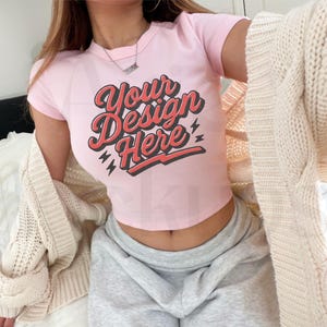 May include: A light pink crop top with the text "Your Design Here" in a retro font. The text is in red with a black outline and lightning bolt accents. The top is short-sleeved.