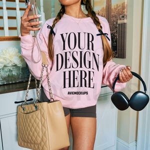 May include: A pink sweatshirt with the text "Your design here" and the words "Aviemockups" printed on the front. The sweatshirt is being worn by a person with a brown leather handbag and black headphones.