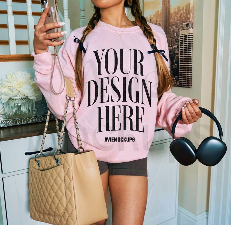 May include: A pink sweatshirt with the text "Your design here" and the words "Aviemockups" printed on the front. The sweatshirt is being worn by a person with a brown leather handbag and black headphones.