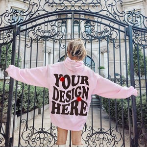 May include: A pink hoodie with the text "YOUR DESIGN HERE" in black lettering with red stars on either side of the text. The hoodie is being worn by a person standing in front of a black metal gate.