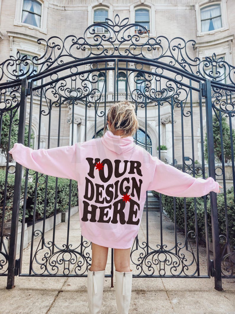 May include: A pink hoodie with the text "YOUR DESIGN HERE" in black lettering with red stars on either side of the text. The hoodie is being worn by a person standing in front of a black metal gate.