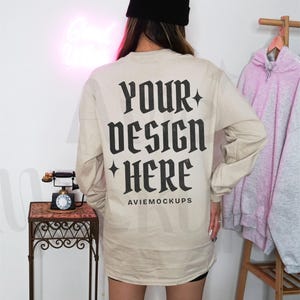 May include: A beige long-sleeved t-shirt with the text "YOUR DESIGN HERE" in black on the back. The text is in a gothic font style. The shirt is being modelled by a person wearing a black beanie.