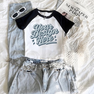 May include: A casual outfit featuring a white and black baseball tee with the text "Your Design Here", light wash jeans, a silver chain belt, white sunglasses, and a mesh tote bag. A newspaper is also included.