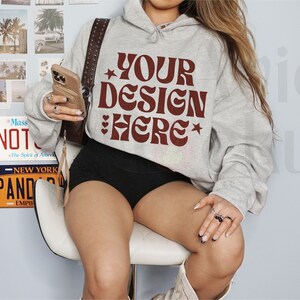 Pu&ograve; includere: Una felpa grigio chiaro con la scritta "YOUR DESIGN HERE" in un font retr&ograve; bordeaux. L'immagine mostra anche pantaloncini neri, una borsa marrone e targhe. La felpa &egrave; in stile casual.