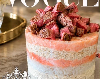 May include: A cylindrical layered food item topped with chunks of cooked red meat. The base consists of alternating horizontal bands of white grains and pinkish-orange textured layers. The text VOGUE appears at the top, and ADORED PETS CUISINE is at the bottom.