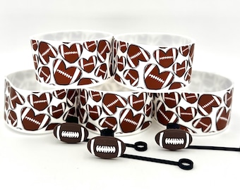 May include: Set of white ceramic bowls and football-shaped bottle stoppers. The bowls feature a repeating pattern of brown footballs in the shape of hearts. The stoppers are brown with white laces and black rubber loops.