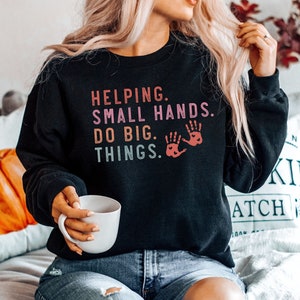 Könnte beinhalten: Schwarzer Pullover mit dem Text "Helping. Small hands do big things." in einem Regenbogen-Farbverlauf. Der Text wird von zwei roten Handabdrücken mit Herzen begleitet.