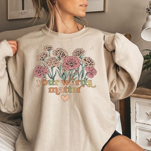 May include: A beige sweatshirt with a floral design and the text "your words matter" in rainbow colors. A small pink heart is below the text.