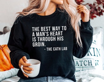Può includere: Felpa nera con testo bianco che recita "THE BEST WAY TO A MAN'S HEART IS THROUGH HIS GROIN. - THE CATH LAB"