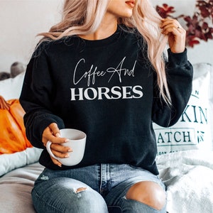 Könnte beinhalten: Ein schwarzer Pullover mit dem Schriftzug "Coffee And Horses" in weißen Buchstaben. Der Pullover wird von einer Person getragen, die eine weiße Tasse hält.