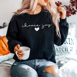 May include: A black sweatshirt with the text "choose joy" in white lettering and a small white heart. The model is wearing blue jeans and holding a white mug.