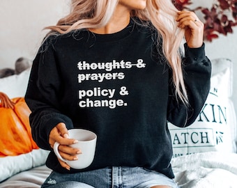 Può includere: Una felpa nera con la scritta "thoughts & prayers policy & change." stampata in bianco sul davanti. La felpa è indossata da una persona con i capelli lunghi e biondi.
