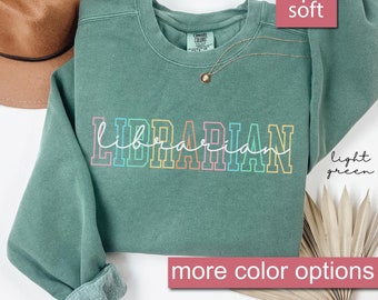 Puede incluir: Una sudadera de color verde claro con la palabra "LIBRARIAN" impresa en colores arcoíris. La sudadera es súper suave y tiene un corte holgado.