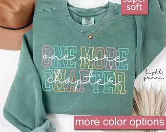 Puede incluir: Sudadera verde claro con el texto "ONE MORE CHAPTER" en letras de bloque coloridas. La sudadera tiene cuello redondo y mangas largas. Un collar de oro con un pequeño colgante está sobre la camisa. También son visibles las palabras "super soft" y "light green".