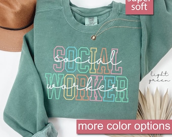 Puede incluir: Una sudadera de color verde claro con el texto "SOCIAL WORKER" en colores arcoíris. La sudadera es súper suave y tiene un corte holgado.
