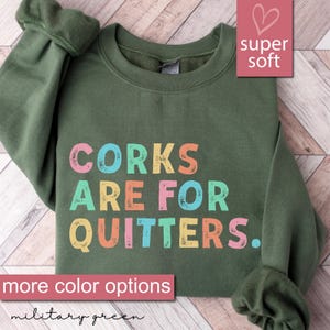 May include: A green sweatshirt with the text "CORKS ARE FOR QUITTERS." in rainbow colors. The sweatshirt is described as "super soft" and "more color options".