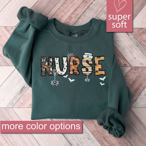 May include: A dark green sweatshirt with the word "NURSE" in colorful Halloween-themed letters. The letters are decorated with spiders, bats, and other Halloween imagery. The sweatshirt is super soft and has more color options.