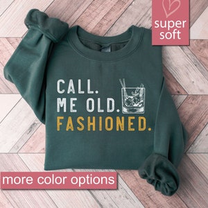 May include: A dark green sweatshirt with the text "CALL. ME OLD. FASHIONED." in white and yellow. The sweatshirt features a graphic of a glass of old fashioned with ice and a cherry.