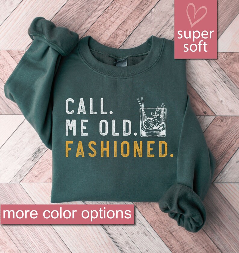 May include: A dark green sweatshirt with the text "CALL. ME OLD. FASHIONED." in white and yellow. The sweatshirt features a graphic of a glass of old fashioned with ice and a cherry.