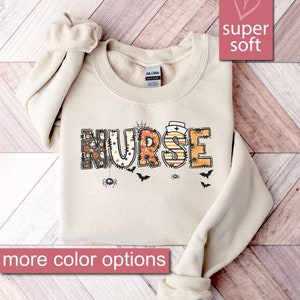 May include: A beige sweatshirt with the word "NURSE" in colorful patchwork letters with a Halloween theme. The letters are decorated with spiders, bats, and ghosts. The sweatshirt is super soft.