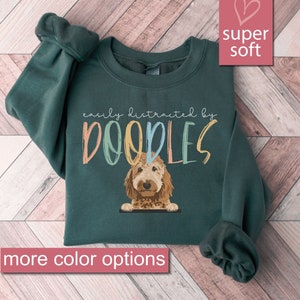 May include: A dark green sweatshirt with the text "easily distracted by doodles" in colorful letters. A doodle dog is peeking over the top of the text. The sweatshirt is super soft.