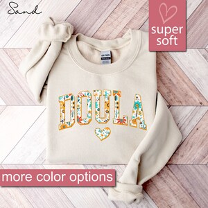 May include: A sand-colored sweatshirt with the word "DOULA" printed in large, yellow letters outlined in black. The letters are filled with a floral pattern in shades of yellow, red, blue, and green. A small heart with a floral pattern is below the word. The text "super soft" is printed in the top right corner of the image.