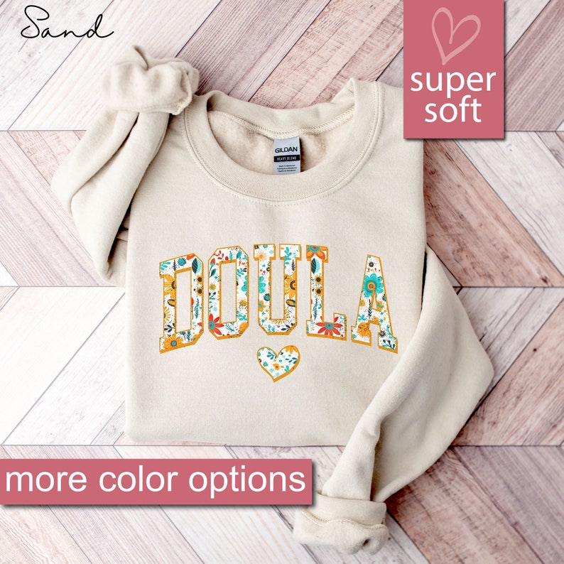 May include: A sand-colored sweatshirt with the word "DOULA" printed in large, yellow letters outlined in black. The letters are filled with a floral pattern in shades of yellow, red, blue, and green. A small heart with a floral pattern is below the word. The text "super soft" is printed in the top right corner of the image.