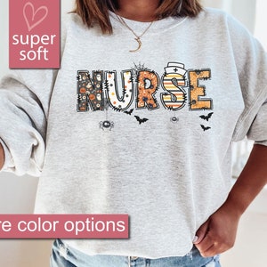 May include: A gray sweatshirt with the word "NURSE" in black, orange, and white letters. The letters are decorated with Halloween-themed patterns, including spiders, bats, and pumpkins.