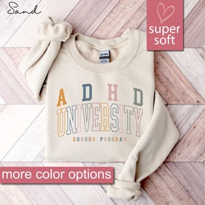May include: A beige sweatshirt with the text "ADHD University Honors Program" in colorful letters. The sweatshirt is on a wood background.