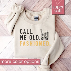 May include: A beige sweatshirt with the text "CALL. ME OLD. FASHIONED." in black and yellow. A drawing of an old fashioned cocktail is next to the text. The sweatshirt is on a wooden surface.
