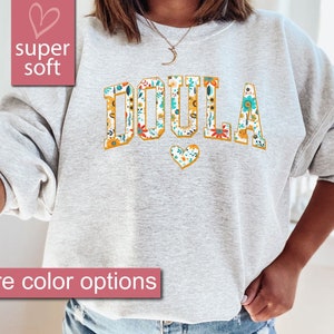 May include: A gray sweatshirt with the word "DOULA" printed in large, yellow letters with a floral pattern. The letters are outlined in gold. A small heart with a floral pattern is below the word.