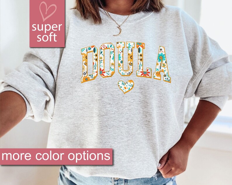 May include: A gray sweatshirt with the word "DOULA" printed in large, yellow letters with a floral pattern. The letters are outlined in gold. A small heart with a floral pattern is below the word.