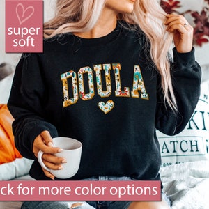 May include: Black sweatshirt with the word "DOULA" in white letters outlined in yellow. The letters are covered in a floral pattern. A small heart with a floral pattern is below the word.
