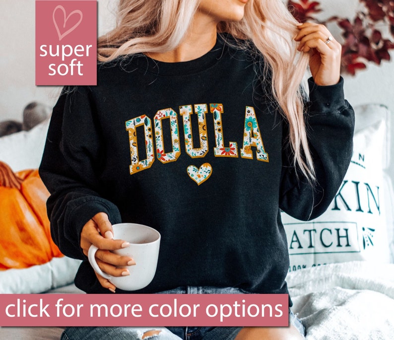 May include: Black sweatshirt with the word "DOULA" in white letters outlined in yellow. The letters are covered in a floral pattern. A small heart with a floral pattern is below the word.